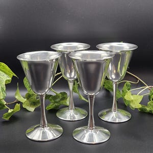 May include: A set of four silver-toned metal goblets with smooth, polished surfaces, flared rims, slender stems, and circular bases. Each goblet features a simple, clean design with a reflective finish, arranged in a group of four identical pieces.