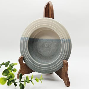 May include: A circular ceramic bowl featuring a two-tone design with a cream-coloured upper section and a speckled blue-grey lower section. The bowl has a series of concentric circular ridges along the rim and is displayed on a wooden stand.