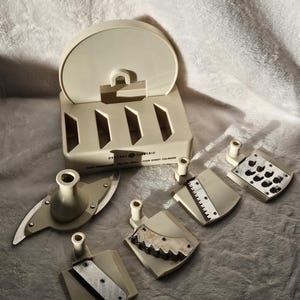 Peut inclure: Robot culinaire General Electric blanc cassé avec plusieurs lames et accessoires. Le robot culinaire a une base rectangulaire avec un dessus incurvé et plusieurs options de lames pour trancher et couper en dés. Le texte "General Electric Pour Robot Culinaire" est visible.
