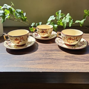 Puede incluir: Tres tazas y platillos vintage con interior crema y ribetes dorados. Las tazas y platillos presentan un diseño floral rojo y dorado. El juego está dispuesto sobre una superficie de madera oscura, con follaje verde al fondo.