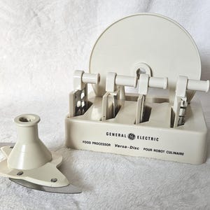 Puede incluir: Una base de almacenamiento para procesador de alimentos General Electric Versa-Disc de color crema con cuatro accesorios de corte metálicos verticales y un conjunto de cuchillas de plástico crema con borde metálico.
