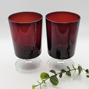 Peut inclure: Deux verres à boire assortis avec des bols rouge foncé translucides, des pieds en verre transparent et des bases circulaires. Les verres présentent une forme cylindrique lisse et sont placés côte à côte, accompagnés d'un petit brin de feuilles vertes.