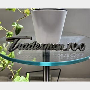 May include: A black, script-style metal emblem featuring the word "Tradesman" followed by the number "100" in a bold, raised font. The piece has a smooth, dark finish and is positioned horizontally, displaying clear, legible lettering throughout.