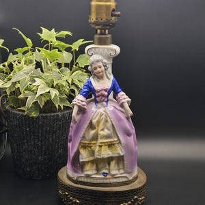 May include: A porcelain table lamp base featuring a figurine of a person in an 18th-century style dress with a blue bodice, pink sleeves, and a lavender skirt. The figure stands on a round, ornate brass-toned metal base with a brass light socket.