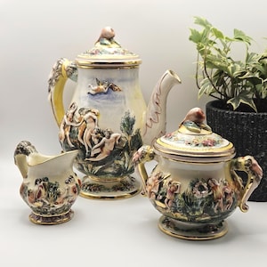 May include: A ceramic tea set consisting of a teapot, a creamer, and a sugar bowl, each featuring raised relief figures of cherubs and mythological scenes in a palette of cream, gold, green, and brown, with fruit-shaped finials on the lids.