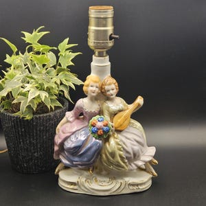 Puede incluir: Una base de lámpara de mesa de porcelana con dos figuras sentadas juntas. Una figura viste un vestido rosa y azul, la otra un vestido amarillo y lavanda, sosteniendo un laúd y un ramo de flores coloridas. La base es blanca con bordes dorados.