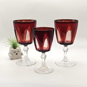 Puede incluir: Tres copas de cristal rojo intenso con tallos y bases transparentes, que presentan un patrón de arco distintivo alrededor del cáliz. Las copas están dispuestas en grupo, junto a una pequeña maceta cuadrada de color gris claro con cara.