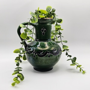 May include: A dark green ceramic pitcher with a glossy finish, featuring a rounded body, a curved handle, and embossed decorative patterns around the neck and midsection, with a trailing green artificial plant draped over the top.