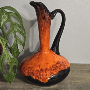 May include: A glass pitcher featuring a bulbous base and a tall, slender neck. The glass displays a vibrant orange colour with a mottled, textured pattern that transitions into a solid, glossy black finish at the base and on the curved handle.