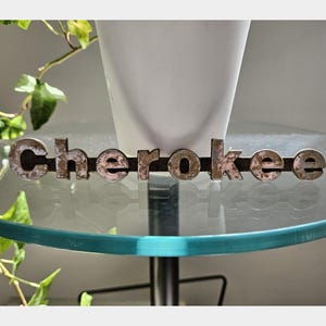 Puede incluir: Un letrero metálico horizontal con la palabra "cherokee" en letras individuales oscuras y desgastadas unidas a una barra de metal delgada. Las letras muestran una superficie texturizada y envejecida con signos visibles de oxidación.
