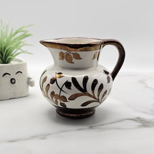 May include: A small ceramic jug with a white base and metallic bronze-coloured leaf and vine patterns. The jug features a rounded body, a curved handle, and a metallic bronze rim. The design includes stylised botanical motifs.