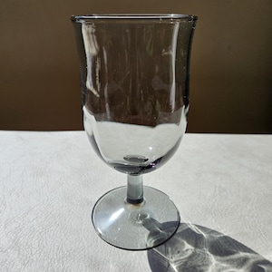 May include: A smoke-colored glass goblet with a smooth, rounded bowl and a short, sturdy stem attached to a circular base. The translucent material features a subtle gray tint, and the design is characterized by its simple, clean, and functional form.