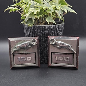 May include: Two square metal emblems featuring a silver-toned leaping ram figure positioned above the number 100. The emblems have a dark, weathered finish with visible signs of wear and oxidation on the edges and surfaces of the metal plates.