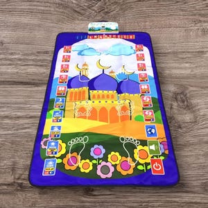 May include: A blue prayer mat with a colorful mosque design. The mat features a central mosque illustration with blue domes and yellow accents. It includes icons for prayer times and a power button.