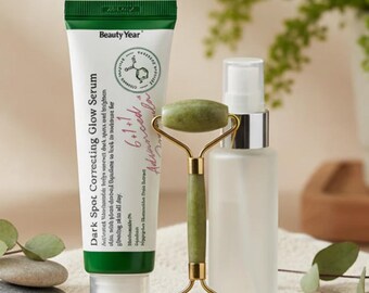 May include: A skincare product arrangement featuring a tube of "Dark Spot Correcting Glow Serum", a jade roller, and a clear spray bottle. The serum tube is white with green accents and text. The jade roller has a gold handle. The items rest on a folded towel.
