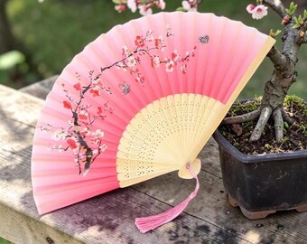 以下が含まれることがあります： ピンク色の扇子。桜と蝶の模様が描かれており、グラデーションのピンク色と明るい木色のベースが特徴です。タッセルが付いています。