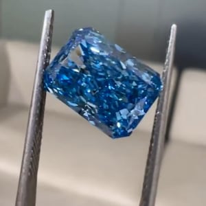 May include: A rectangular cut gemstone with a vibrant, deep blue color and multiple facets that create a high level of brilliance and light reflection. The stone is held by metal tweezers, showcasing its clear, saturated blue hue and sharp edges.