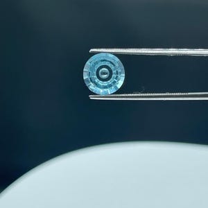 Op de afbeelding: Een ronde, lichtblauwe edelsteen met een gefacetteerd oppervlak en een concentrisch cirkelpatroon, vastgehouden door een zilverkleurige metalen pincet. De steen heeft een helder, gepolijst uiterlijk met duidelijke geometrische sneden vanuit het midden.