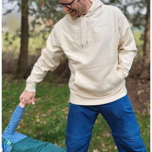May include: A cream-coloured hoodie with a front pocket and drawstring, worn with blue trousers. The hoodie features a small logo on the chest. The person is holding the hand of another person, wearing a blue and white striped top.