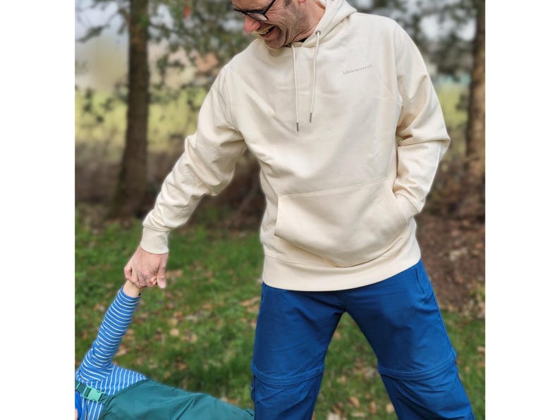 May include: A cream-coloured hoodie with a front pocket and drawstring, worn with blue trousers. The hoodie features a small logo on the chest. The person is holding the hand of another person, wearing a blue and white striped top.