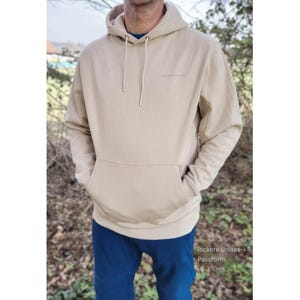 May include: A beige hoodie with a front pocket and a drawstring hood. The hoodie has a small logo on the chest and is worn with blue trousers. The text "lockere Unisex Passform" is visible at the bottom of the image.