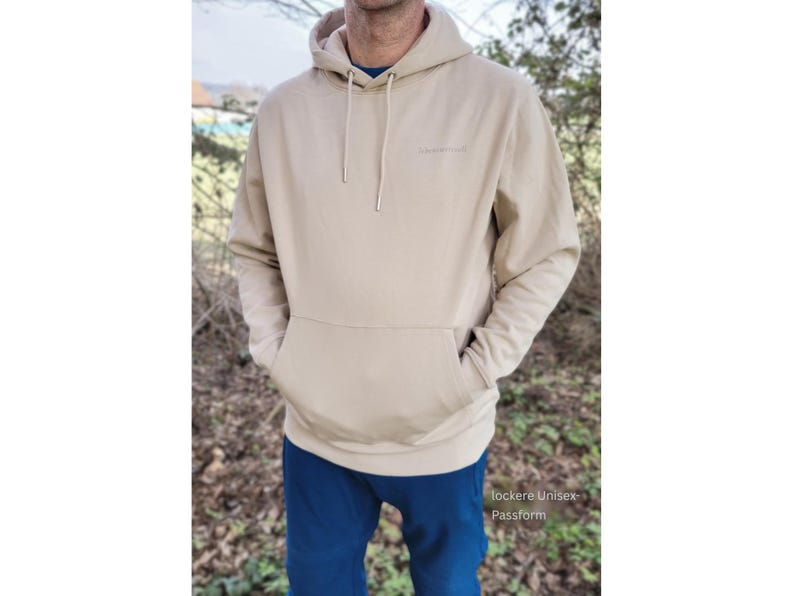 May include: A beige hoodie with a front pocket and a drawstring hood. The hoodie has a small logo on the chest and is worn with blue trousers. The text "lockere Unisex Passform" is visible at the bottom of the image.