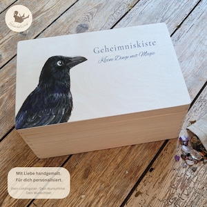 Könnte beinhalten: Eine rechteckige Kiste aus hellem Holz mit einer detaillierten Illustration eines schwarzen Raben auf der linken Seite. Der Text "Geheimniskiste" und "Kleine Dinge mit Magie" ist in Dunkelblau auf den Holzdeckel gedruckt.