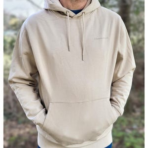 May include: A beige hoodie with a drawstring hood and a front pocket. The word "lebesraum" is printed on the upper left chest. The person is wearing a blue shirt under the hoodie.