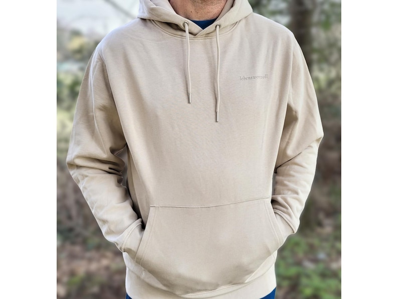 May include: A beige hoodie with a drawstring hood and a front pocket. The word "lebesraum" is printed on the upper left chest. The person is wearing a blue shirt under the hoodie.