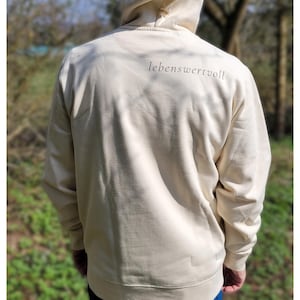 May include: Cream-coloured hoodie with the word "lebenswertvoll" printed in a dark font on the upper back. The hoodie has a drawstring hood and long sleeves. The person is wearing blue jeans.
