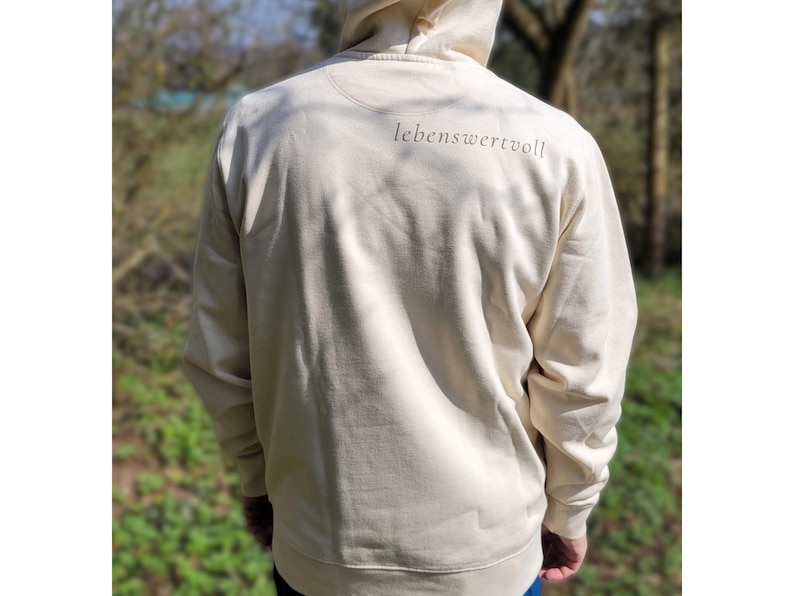 May include: Cream-coloured hoodie with the word "lebenswertvoll" printed in a dark font on the upper back. The hoodie has a drawstring hood and long sleeves. The person is wearing blue jeans.