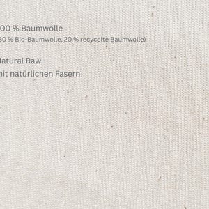 May include: Close-up of a cream-coloured fabric with text. The text reads "100% Baumwolle (80% Bio-Baumwolle, 20% recycelte Baumwolle)", "Natural Raw", and "mit nat&uuml;rlichen Fasern". The fabric has a textured appearance.