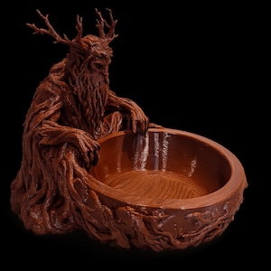 Puede incluir: Un cuenco marrón texturizado que presenta una figura esculpida con astas y una barba que se asemeja a raíces y ramas de árbol. La figura está colocada detrás del cuenco circular, con las manos apoyadas en el borde, creando un diseño orgánico.