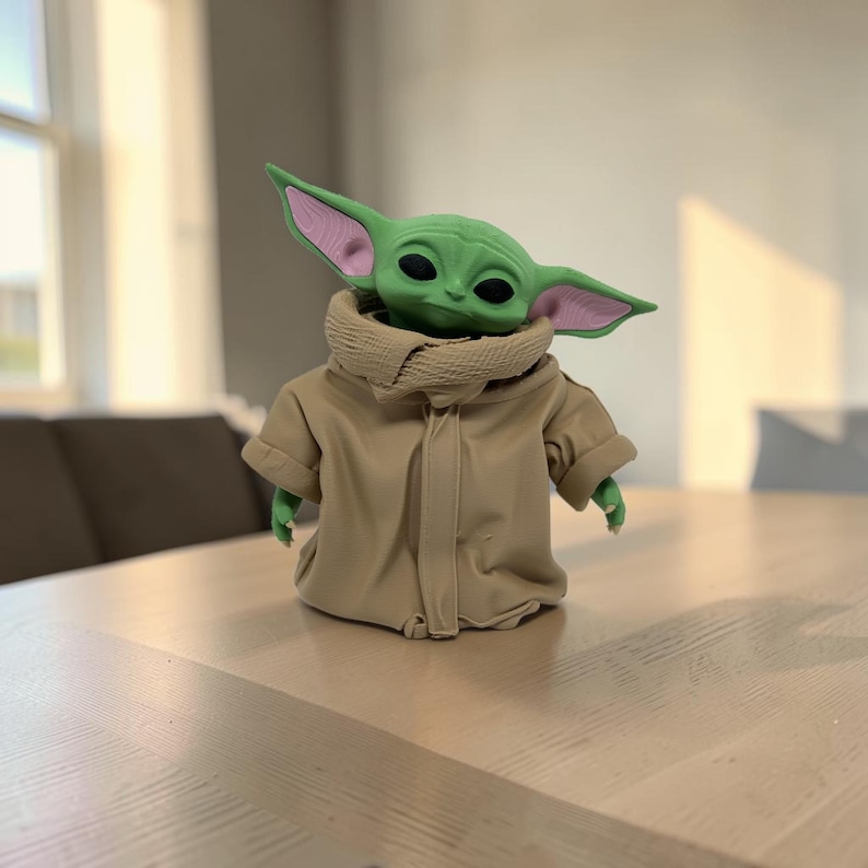 May include: A small green creature figurine with large pointed ears, black eyes, and a textured light pink inner ear detail. The figure is wearing a tan-colored robe with a high, folded collar and long sleeves that extend past the small hands.