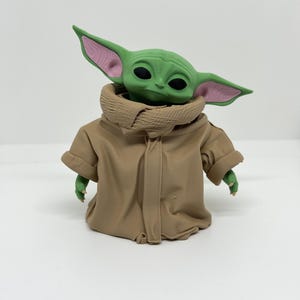 May include: A small green creature figurine with large pointed ears featuring pink inner details and black eyes. The figure is wearing a tan-colored robe with a high, textured collar that wraps around the neck, covering the torso and arms.