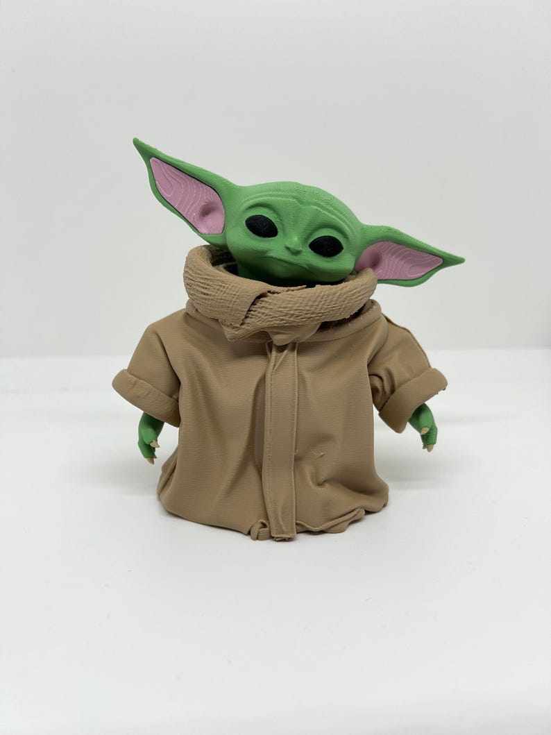 May include: A small green creature figurine with large pointed ears featuring pink inner details and black eyes. The figure is wearing a tan-colored robe with a high, textured collar that wraps around the neck, covering the torso and arms.