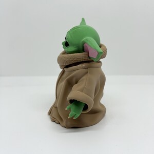 May include: A small green figurine with large ears, black eyes, and a textured surface. The figure wears a tan robe with a high collar and has visible fingers on its small green hands. The character is shown from a side profile view.