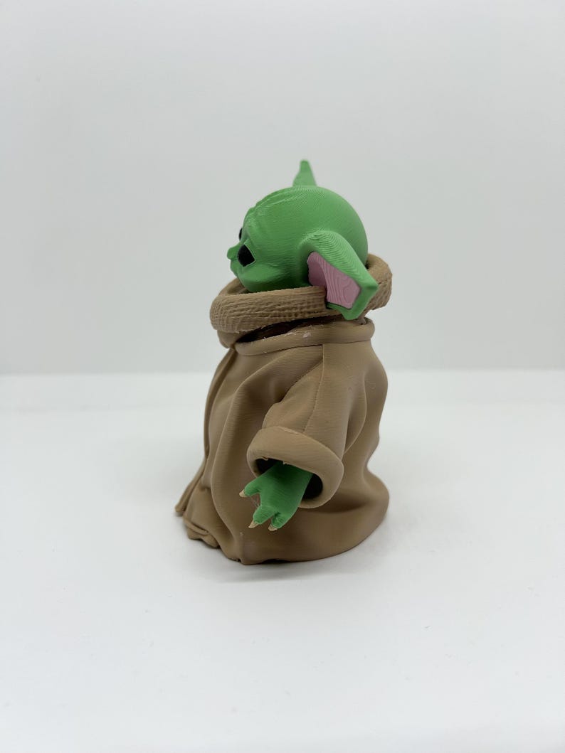 May include: A small green figurine with large ears, black eyes, and a textured surface. The figure wears a tan robe with a high collar and has visible fingers on its small green hands. The character is shown from a side profile view.
