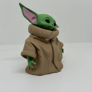 May include: A small green figurine with large pointed ears, dark eyes, and a textured tan robe with a high collar. The character has a smooth green head with pink inner ear details and small hands extending from the sleeves of the tan garment.