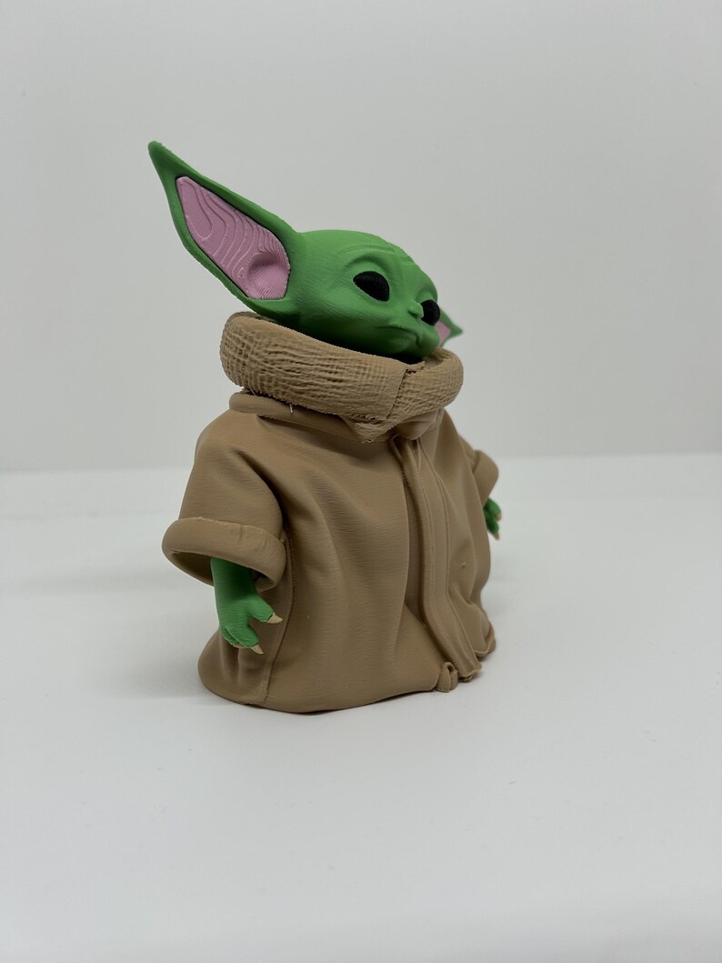 May include: A small green figurine with large pointed ears, dark eyes, and a textured tan robe with a high collar. The character has a smooth green head with pink inner ear details and small hands extending from the sleeves of the tan garment.