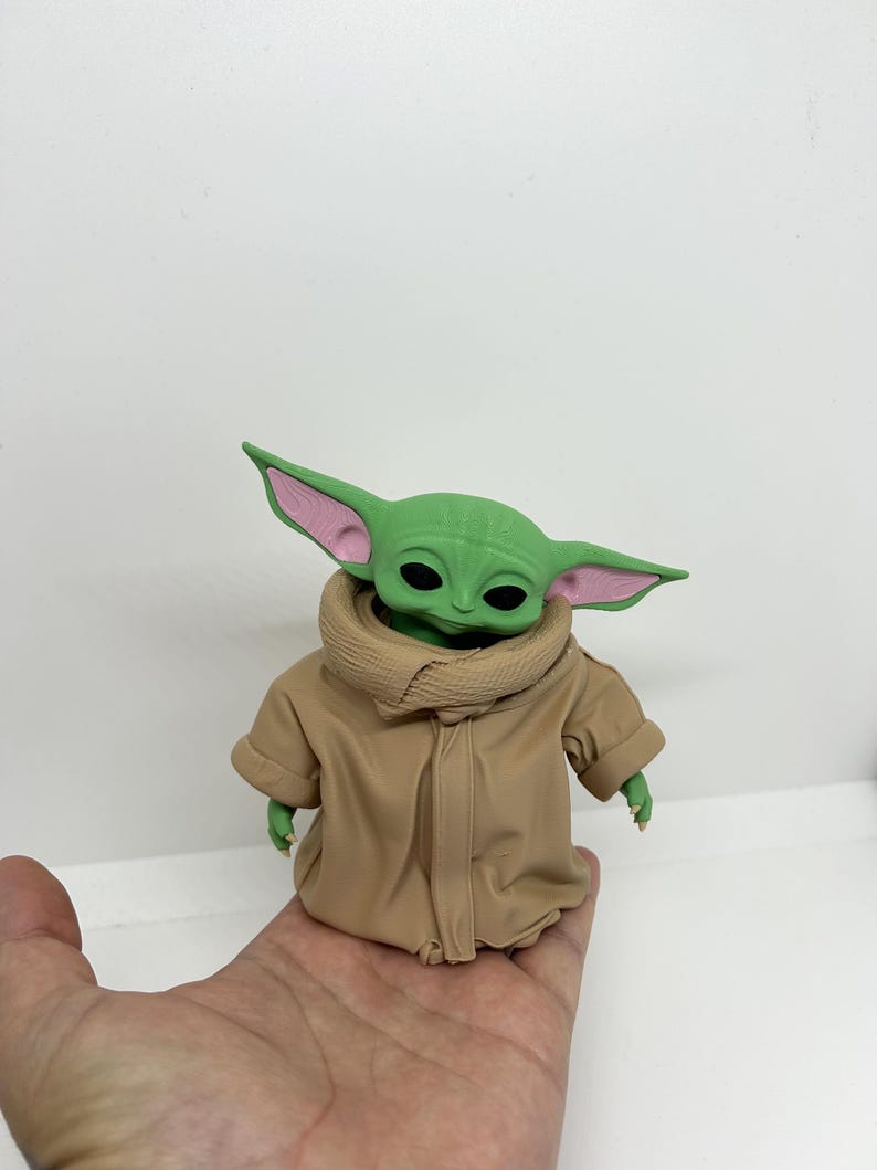 May include: A small green creature figurine with large pointed ears, black eyes, and a light pink inner ear detail. The figure is wearing a tan-colored robe with a high, textured collar that wraps around the neck and covers the torso.