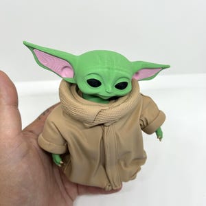 May include: A small green creature figurine with large pointed ears, black eyes, and a textured head surface. The figure wears a tan-colored robe with a high, folded collar and visible fabric folds, featuring pink accents inside the ears.