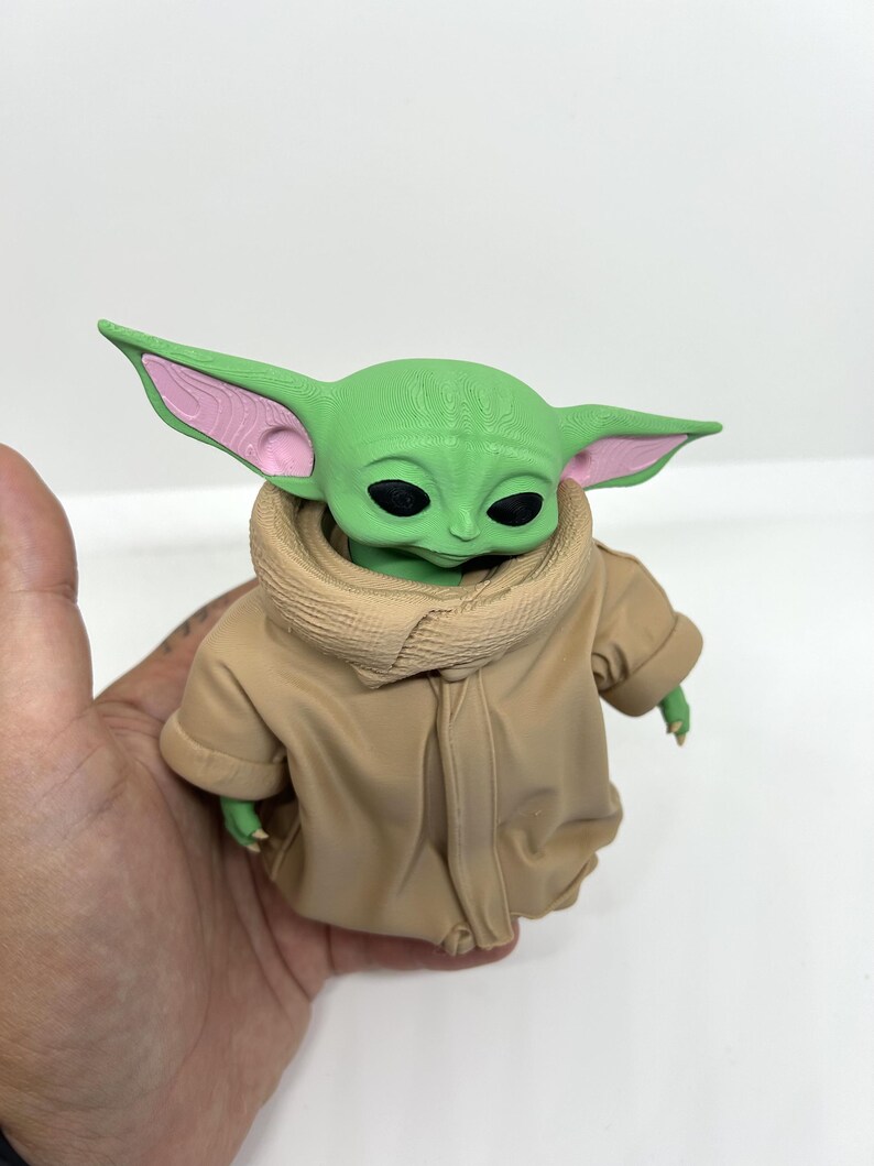 May include: A small green creature figurine with large pointed ears, black eyes, and a textured head surface. The figure wears a tan-colored robe with a high, folded collar and visible fabric folds, featuring pink accents inside the ears.