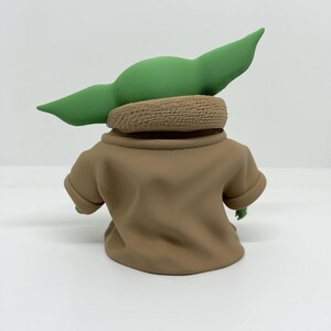 May include: A small figurine of a green creature with large pointed ears, wearing a tan robe with a textured collar. The figure is viewed from the back, showing the smooth matte finish of the garment and the distinct shape of the head and ears.