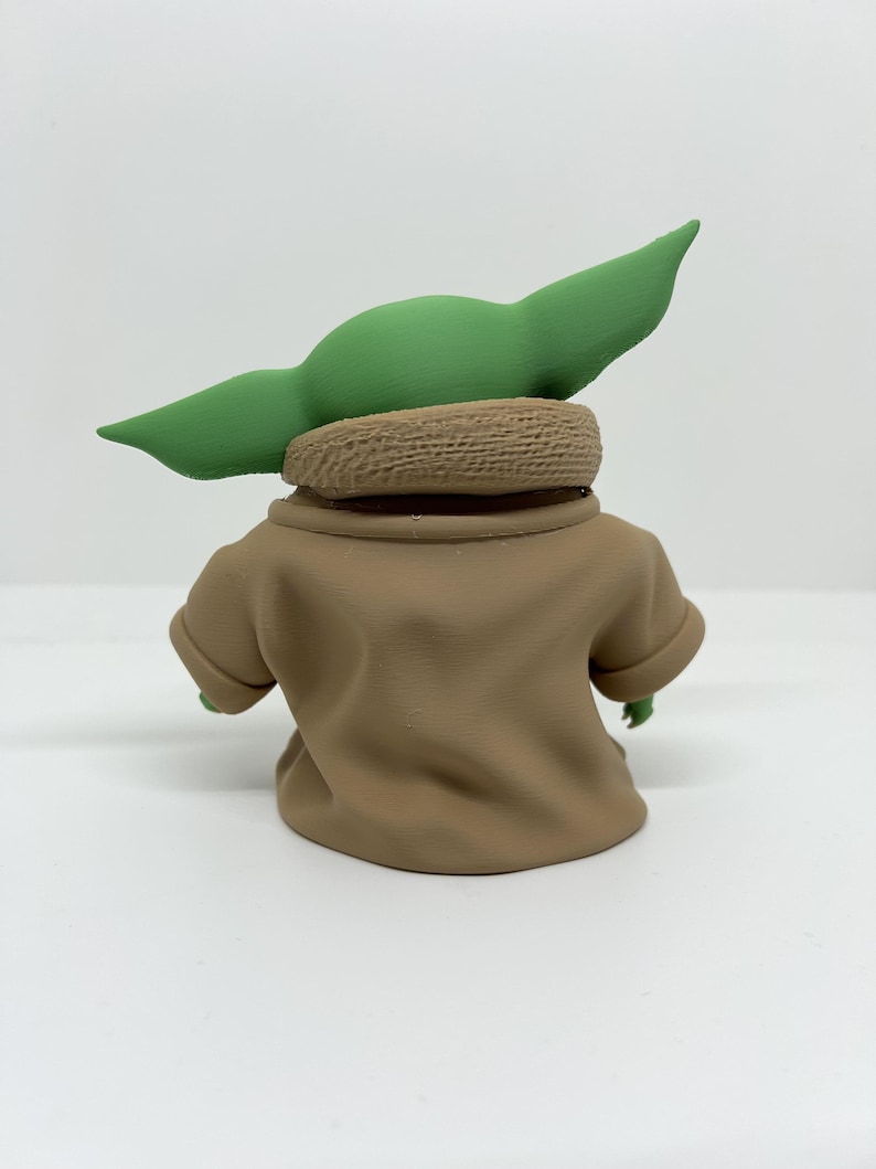 May include: A small figurine of a green creature with large pointed ears, wearing a tan robe with a textured collar. The figure is viewed from the back, showing the smooth matte finish of the garment and the distinct shape of the head and ears.