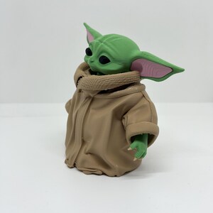 May include: A small green creature figurine with large pointed ears, black eyes, and pink inner ear details. The figure is wearing a tan-colored robe with a textured collar and wide sleeves, showing small green hands with tiny claws extending from the garment.