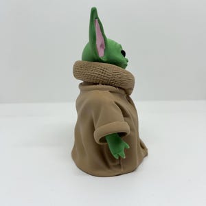 May include: A green-skinned figurine with large pointed ears featuring pink inner details. The character has a smooth head and is wearing a tan-colored robe with a textured, thick collar that wraps around the neck and shoulders.