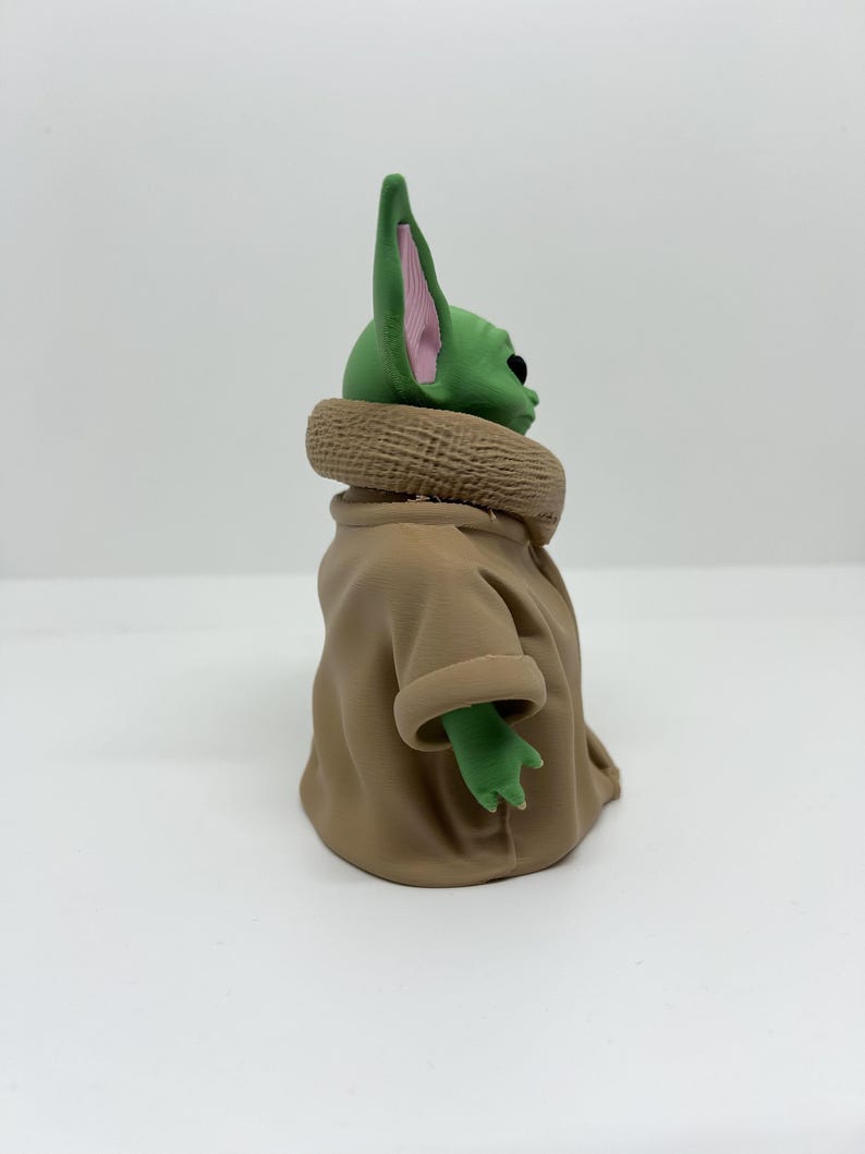 May include: A green-skinned figurine with large pointed ears featuring pink inner details. The character has a smooth head and is wearing a tan-colored robe with a textured, thick collar that wraps around the neck and shoulders.