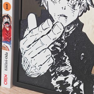 May include: Black and white manga-style art piece featuring a character with a hand gesture. The artwork is framed in black and stands next to several manga books with colorful covers. The books are labeled with the number "1".