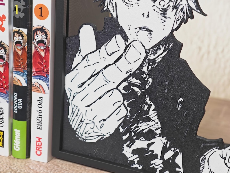 May include: Black and white manga-style art piece featuring a character with a hand gesture. The artwork is framed in black and stands next to several manga books with colorful covers. The books are labeled with the number "1".